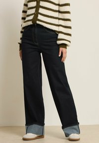 Tummat denim leveät housut, joissa on vaaleansiniset käänteet, yhdistettynä vihreään ja kermaiseen raidalliseen neulecardiganiin, jossa on pyöreät napit.