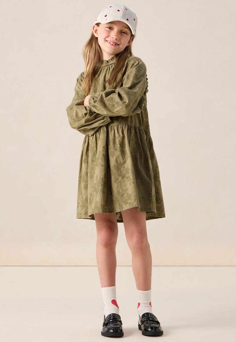 Mädchen, das lächelt und die Arme verschränkt, trägt ein olivgrünes Langarmkleid, eine weiße Mütze mit roten Herzen, weiße Socken und schwarze Slipper vor einem beigen Hintergrund.