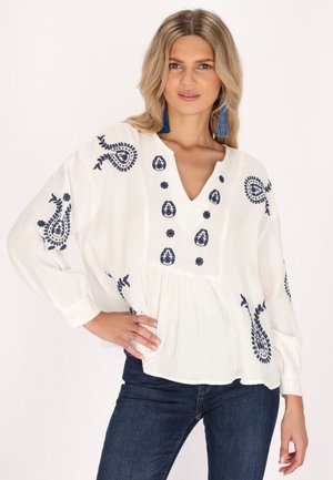 White blouse with navy blue embroidery in paisley and floral patterns, V-neckline, long sleeves, and a loose fit, paired with dark jeans.