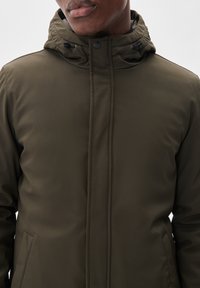 Man som bär en olivgrön vinterjacka med huva, dragkedja framtill och snörning vid nacken.