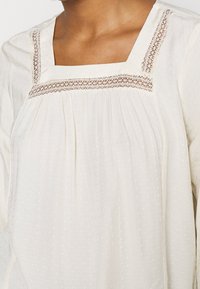 Weißes Blusenshirt mit quadratischem Ausschnitt, aufwendiger Spitzenverzierung entlang des Ausschnitts und weichem, strukturiertem Stoff mit dezenten Punkten.