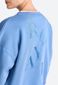 Light blue sweatshirt featuring a textured, embroidered design spelling "ROYAL" on the back. Round neckline and relaxed fit.