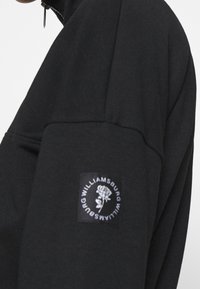 Svart sweatshirt med hög krage, dragkedja och ett märke med vit text och en rosdesign på vänster ärm.