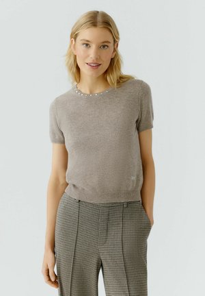 Beige Strickoberteil mit kurzen Ärmeln und strukturierter Oberfläche, versehen mit einem runden Ausschnitt, der mit kleinen dekorativen Perlen verziert ist. Trägt dazu gemusterte Hosen.