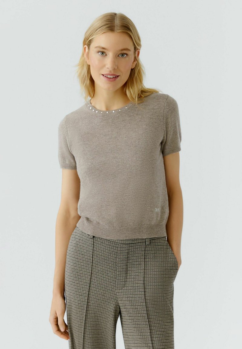 Beige Strickoberteil mit kurzen Ärmeln und strukturierter Oberfläche, versehen mit einem runden Ausschnitt, der mit kleinen dekorativen Perlen verziert ist. Trägt dazu gemusterte Hosen.