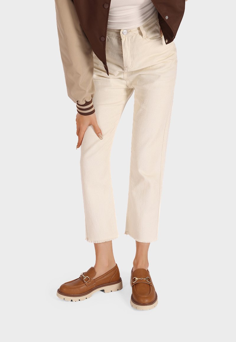 Mocassini in pelle color tan con hardware dorato, pantaloni cream a vita alta con orlo sfilacciato, giacca marrone a strati con polsini a costine, top bianco.