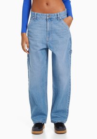 Osoba nosząca jasnoniebieskie, szerokie jeansy z wysokim stanem, z rękami w kieszeniach i czarnymi trampkami z gumowymi podeszwami, skrócone powyżej talii.