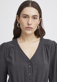 Schwarze Bluse mit V-Ausschnitt, puffigen Schultern und sechs Knöpfen auf der Vorderseite. Glattes Material mit leichtem Glanz und taillierter Passform.