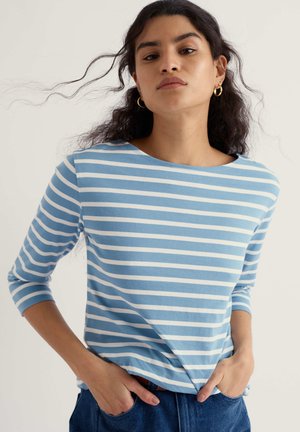 Ženska s kodrastimi lasmi, ki nosi zlate obročaste uhane in modro-belo črtasto majico z dolgimi rokavi, z rokami v žepih hlač.