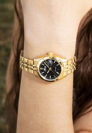 Montre dorée avec cadran noir, affichage de la date et bracelet en métal. Boîtier rond avec couronne texturée, dotée d'un mouvement à quartz.