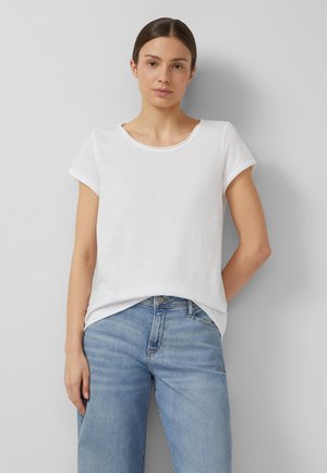 Frau trägt ein schlichtes weißes kurzärmeliges T-Shirt, leicht in hellblaue Jeans gesteckt, steht vor einem hellgrauen Hintergrund.