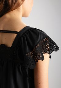 Top nero con maniche corte a volant e dettagli in croché intricati. Tessuto morbido, vestibilità rilassata e design con spalline posteriori.