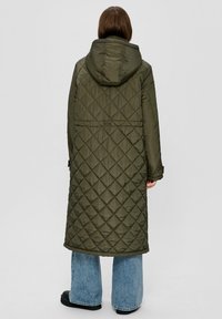 Olijfgroene, gewatteerde lange jas met een capuchon, voorzien van ruitenpatronen en een rechte snit, gecombineerd met blauwe wijde jeans.