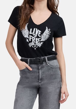 Kobieta ubrana w czarny T-shirt z dekoltem w serek z napisem "Live to Free", skrzydłami i wzorem szachownic, w zestawie ze szarymi dżinsami i czarnym paskiem z ćwiekami.