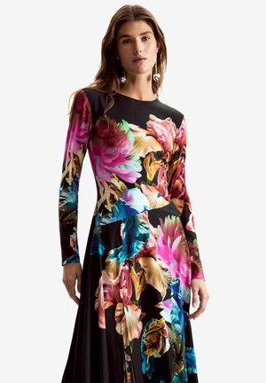 Kobieta w długiej czarnej sukience z dużym kolorowym kwiatowym wzorem w różowym, niebieskim, żółtym i zielonym kolorze, pozująca na tle jednolitego tła.