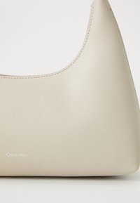 Bež torba od umjetne kože s glatkom teksturom, zaobljenim dizajnom i suptilnim šavovima. Ima utisnut "Calvin Klein" logo na prednjoj strani.