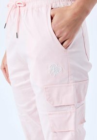 Helle rosa Cargo-Hosen mit einem elastischen Bund, Kordelzug und mehreren Taschen. Mit einem weißen gestickten Logo auf der Vorderseite.