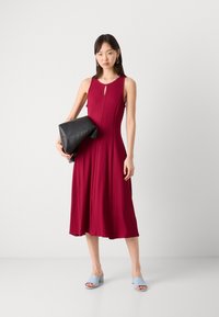 Punainen hihaton mekko, jossa on avainaukko yksityiskohtana ja liehuva siluetti. Yhdistetty vaaleansinisiin liukuvarsikonjokiin ja mustaan clutch-laukkuun.