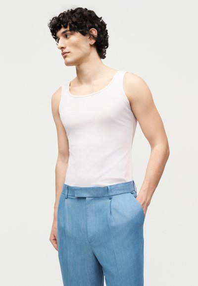 Homme aux cheveux bouclés portant un débardeur blanc et un pantalon bleu clair, debout, une main dans la poche, regardant vers la gauche.
