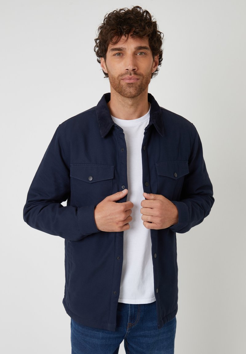Granatowa koszula-kurtka z guzikami, z miękkim kołnierzykiem z sztruksu. Posiada dwie kieszenie na piersi oraz luźny krój, noszona na białej koszulce.