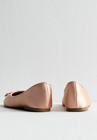 Rosaguld satinballerina med rund tå, detalj bak och platt gummisula. Inkluderar en dekorativ rosett fram.