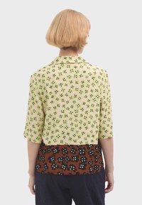 Korte mouw blouse met een lichtgroen bloemenpatroon op beige stof, met een contrasterend donkerbruin bloemenontwerp aan de zoom.