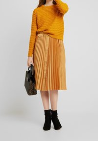 Pull en tricot moutarde, jupe plissée beige avec boutons, bottines noires et sac noir avec un motif de texture serpent.