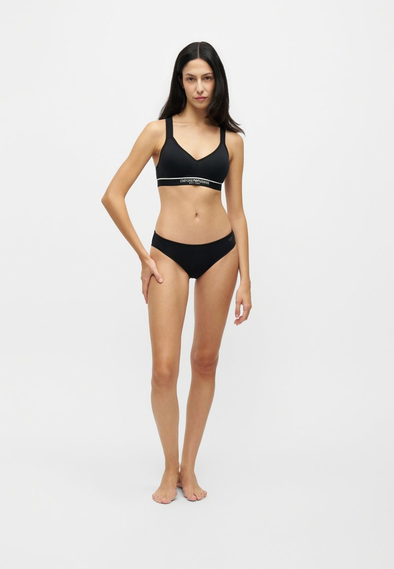 Femme debout pieds nus, portant un soutien-gorge noir et une culotte noire assortie sur un fond clair uni.