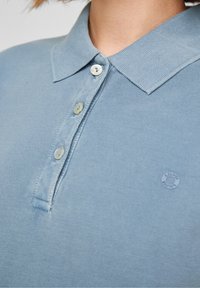 Vaaleansininen poolopaita, joka on valmistettu teksturoidusta puuvillasta. Paidassa on klassinen kaulus, viisi nappia ja pieni logo yksityiskohta oikeassa alakulmassa.