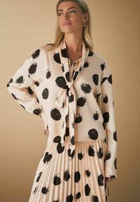 Rosa bluse med sorte penselstrøg polka prikker, lange ærmer, en sløjfe ved halsen, parret med en plisseret nederdel med matchende mønstre.