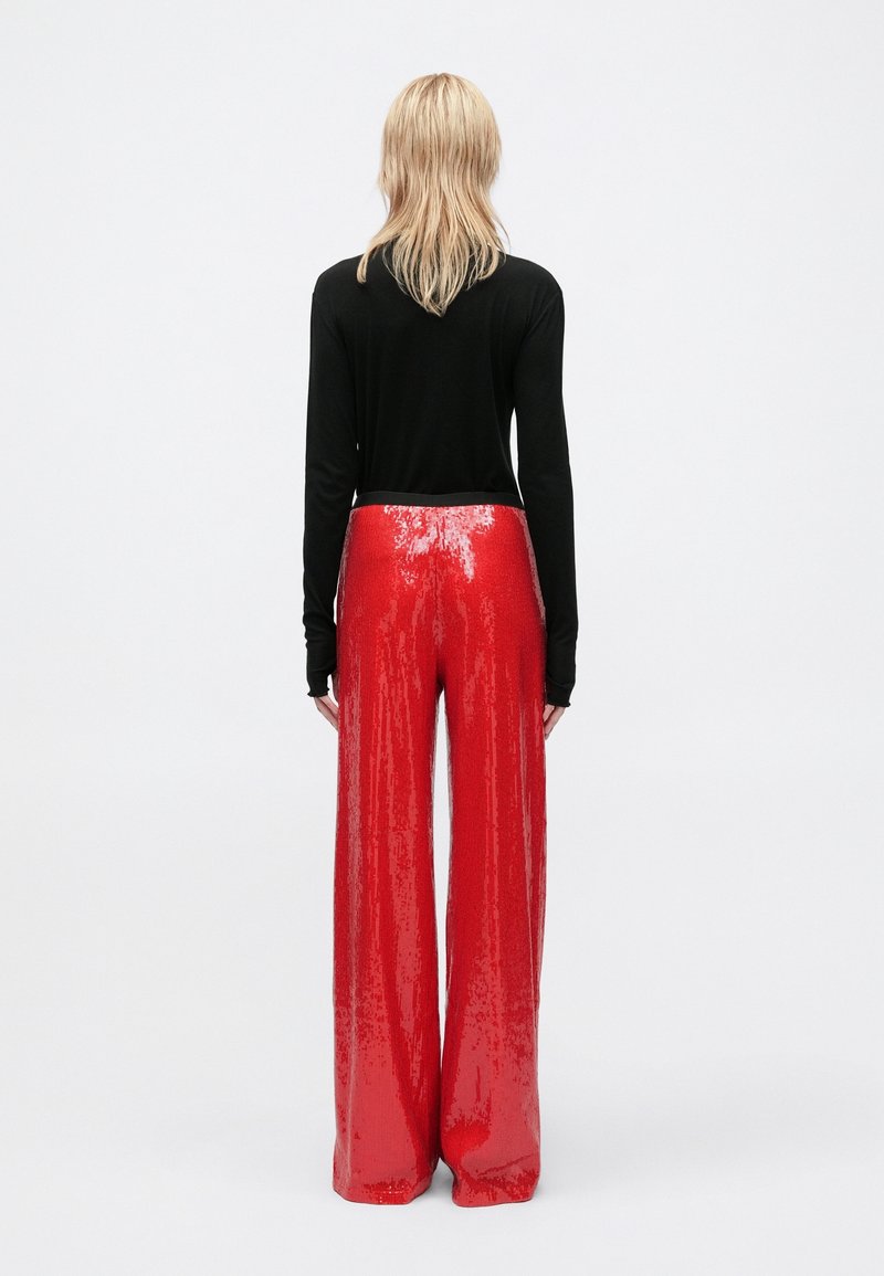 Personne debout de dos, portant un haut noir à manches longues et un pantalon rouge brillant à paillettes, taille haute et large, devant un fond uni.