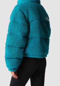 Tyrkysová fleecová bunda s texturovaným, chlupatým povrchem. Má krátký střih a vyšitý logo na zádech, kombinovaná s černými kalhotami.