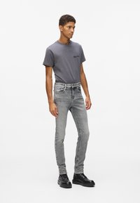 Pelēks kokvilnas T-krekls ar īsām piedurknēm un mazu logotipu, apvienots ar slaidām gaismas pelēkām džinsu biksēm un melniem aukliņu potīšu zābakiem.