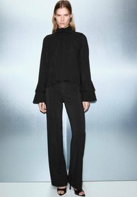 Črna bluza z dolgimi rokavi in visokim ovratnikom, z nabranim dizajnom, v kombinaciji z ustreznimi širokimi hlačami. Slikovita tkanina in razširjeni manšeti.