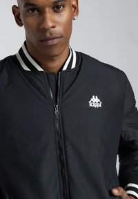 Giubbotto bomber nero con colletto a righe bianche, chiusura con zip e logo Kappa ricamato sul petto sinistro. Materiale morbido e leggero.