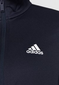 Zbliżenie na czarną sportową kurtkę zapinaną na zamek, z białym logo Adidas na piersi.