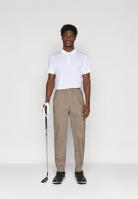 Tricou polo alb cu guler, pantaloni bej, pantofi de golf negri și o rachetă de golf; modelul stă drept cu un mănuș pe mâna stângă.