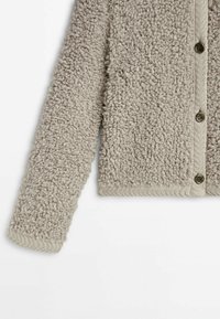 Cardigan beige texturé avec un extérieur duveteux, des poignets côtelés et des boutons le long du devant avec une patte tricotée.