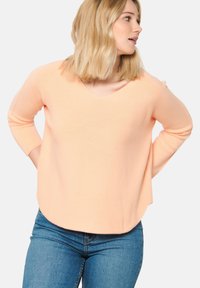 Maglione a maglia color pesca con scollo a V, maniche lunghe e vestibilità comoda. Tessuto liscio e orlo arrotondato, abbinato a jeans blu.