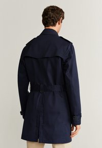 Mężczyzna ubrany w granatowy płaszcz trencz z paskiem i detalami z klamrami, stoi tyłem na tle jednolitego jasnego tła.