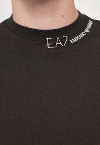 Fekete póló bordázott kerek nyakkal. A bal felső részen fehér hímzett "EA7" és "EMPORIO ARMANI" felirat található. Simaságot biztosító textúra.