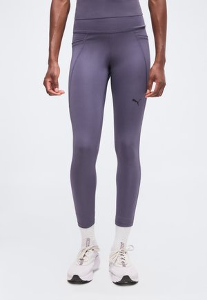 Personne portant des leggings moulants violets, des chaussettes blanches et des baskets blanches Puma, debout devant un fond uni.