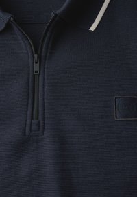 Tmavě modrý svetr s límcem na zip, vyrobený z texturované látky, s malou černou nášivkou a bílým pruhom okolo límce.