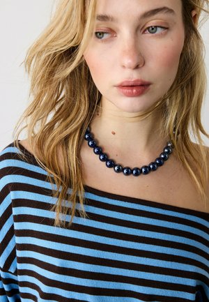 Młoda kobieta o falowanych blond włosach, ubrana w niebiesko-czarną bluzkę z odkrytymi ramionami i ciemnoniebieski naszyjnik z koralików.