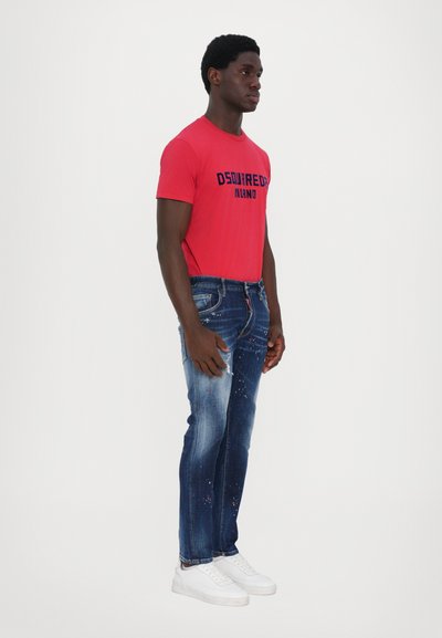 Camiseta ajustada roja con "DSQUARED2 MILANO" impresa en negro, combinada con jeans azules desgastados con salpicaduras de pintura y zapatillas blancas.