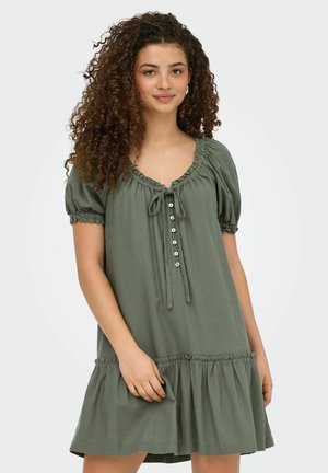 Ung kvinde med krøllet hår iført en løs olivengrøn kjole med pufærmer og knapdetaljer, stående mod en ensfarvet baggrund.