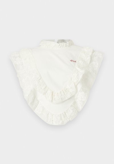 Λευκό μπλουζάκι με ψηλό γιακά και φαρδιά δαντελένια βολάν κατά μήκος των ώμων και του στήθους, με μικρό κόκκινο κεντητό κείμενο στο επάνω μέρος του στήθους.