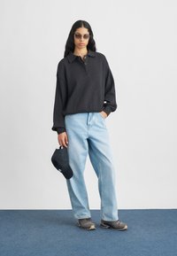 Mulher vestindo um suéter escuro oversized, jeans claros azuis, tênis cinzentos, óculos de sol, segurando um boné preto, em pé sobre um tapete azul contra uma parede branca.