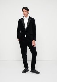 Melnas samta fraka ar satīna apkakli, kopā ar melna samta biksēm un baltu uzvalka krekli. Modelis valkā melnas kurpes.