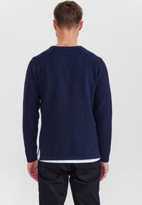 Mörkblå texturerad stickad tröja med långärmar och rund hals; sidospalt detaljer; bärs över en vit skjorta; visas bakifrån.